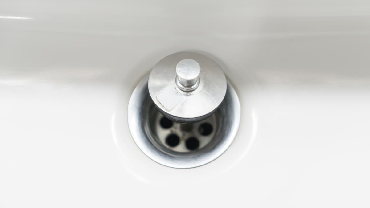 Desentupidor de borracha vermelho apoiado sobre o ralo de uma pia de cozinha de inox com água parada