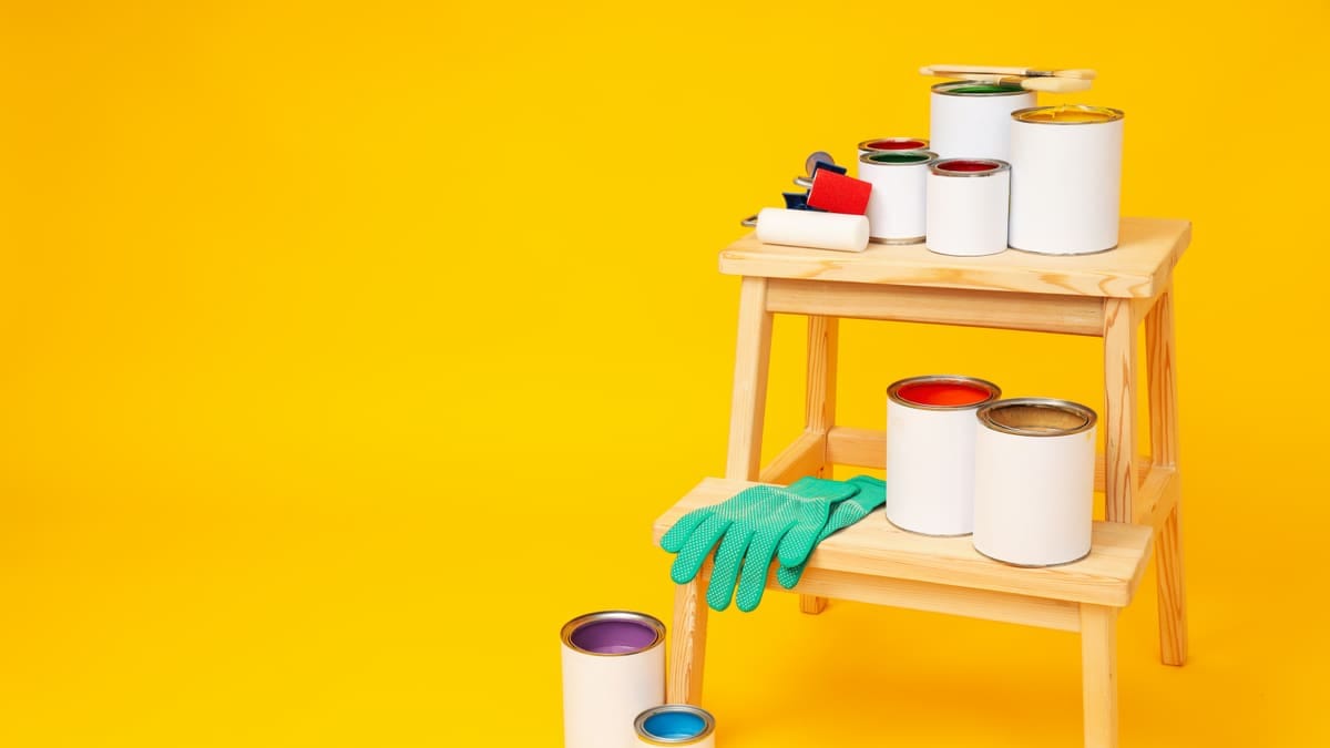 Latas de tinta abertas com rolo e bandeja sobre lona, ao lado de uma parede pintada pela metade